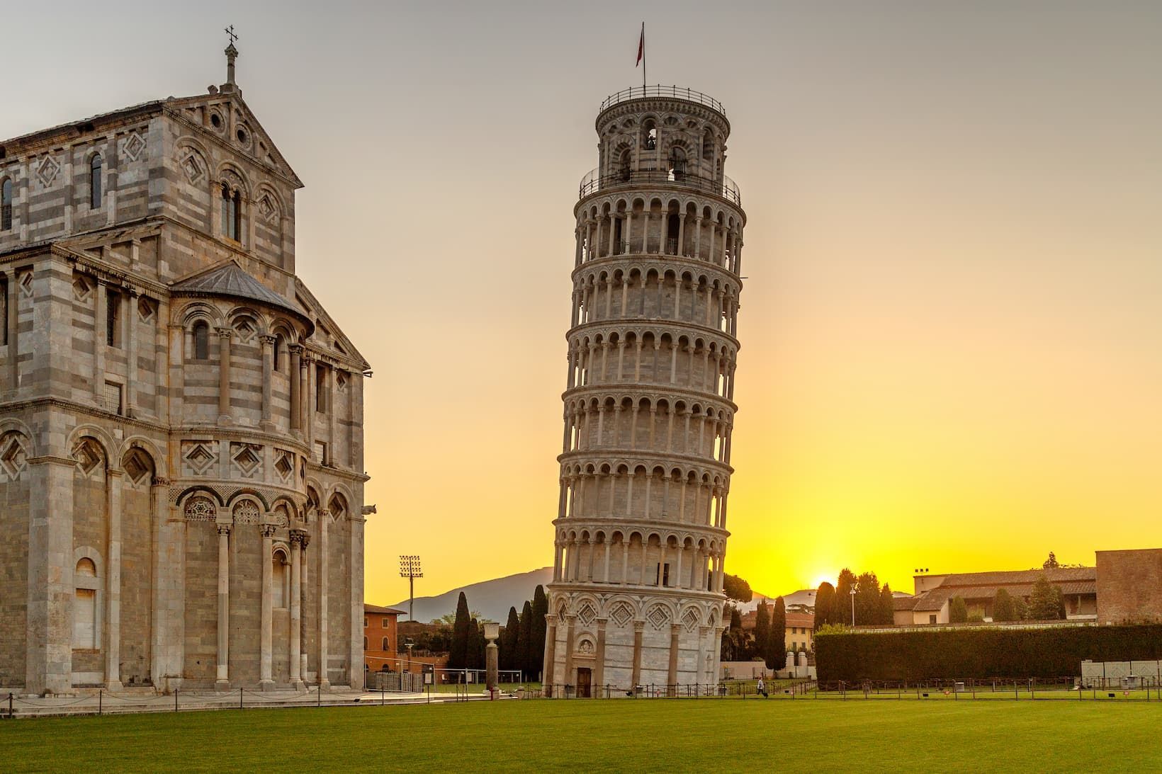 A leaning bell tower and an ornate cathedral stand on a green lawn against a golden sunset.