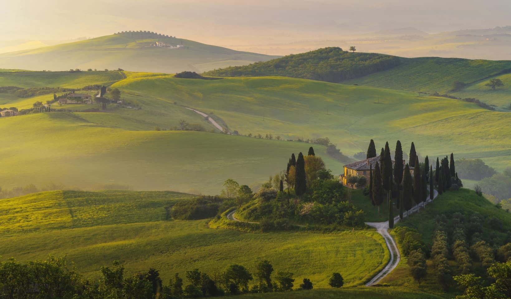 A villa surrounded by cypress trees sits on a rolling green hill amidst a hazy landscape at sunrise.