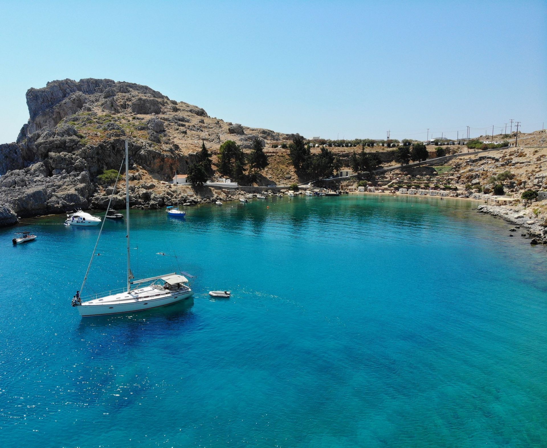 Una barca a vela bianca ancorata in una baia di acqua turchese, con una costa rocciosa e una piccola spiaggia sullo sfondo.
