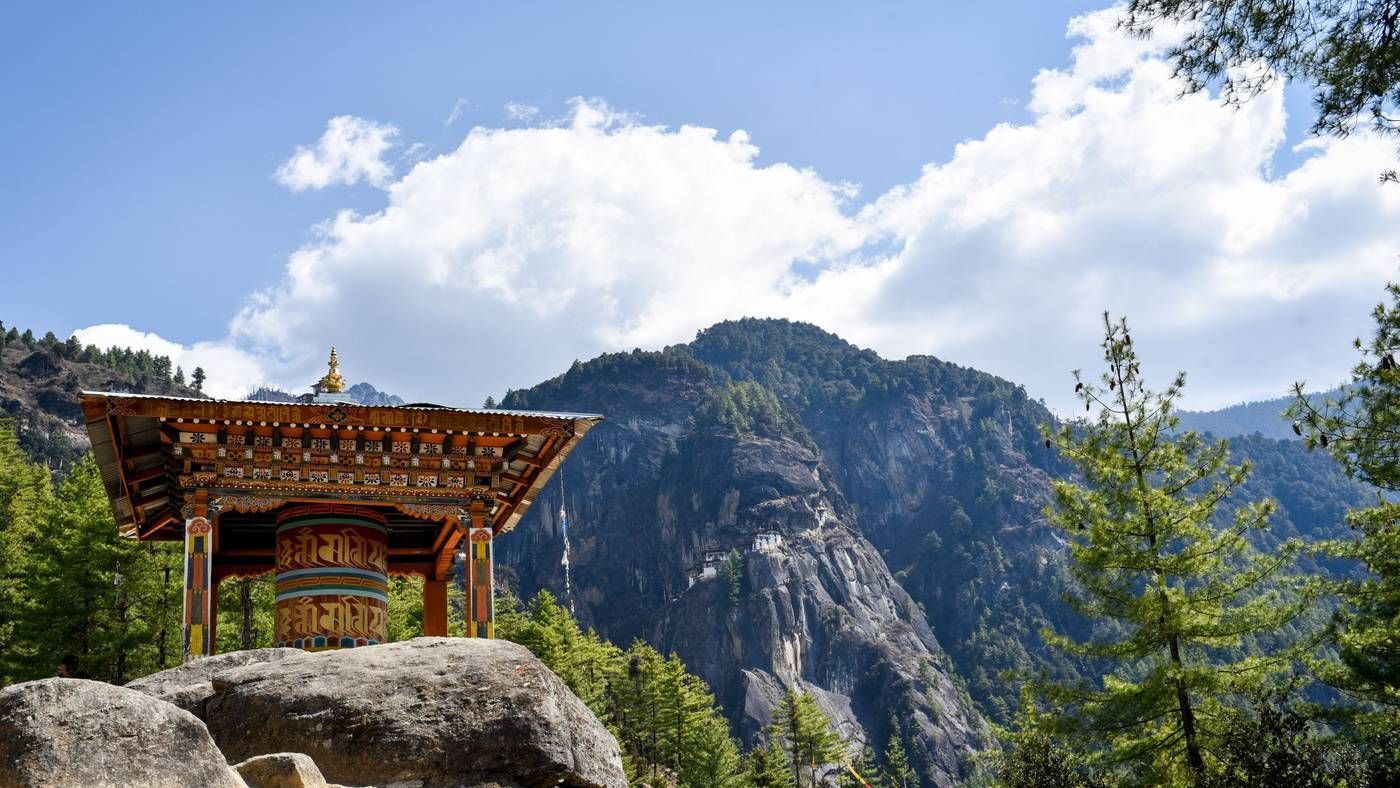 Una ruota di preghiera colorata in un padiglione di legno su una roccia, che si affaccia su una montagna boscosa con un monastero sulla parete della scogliera.