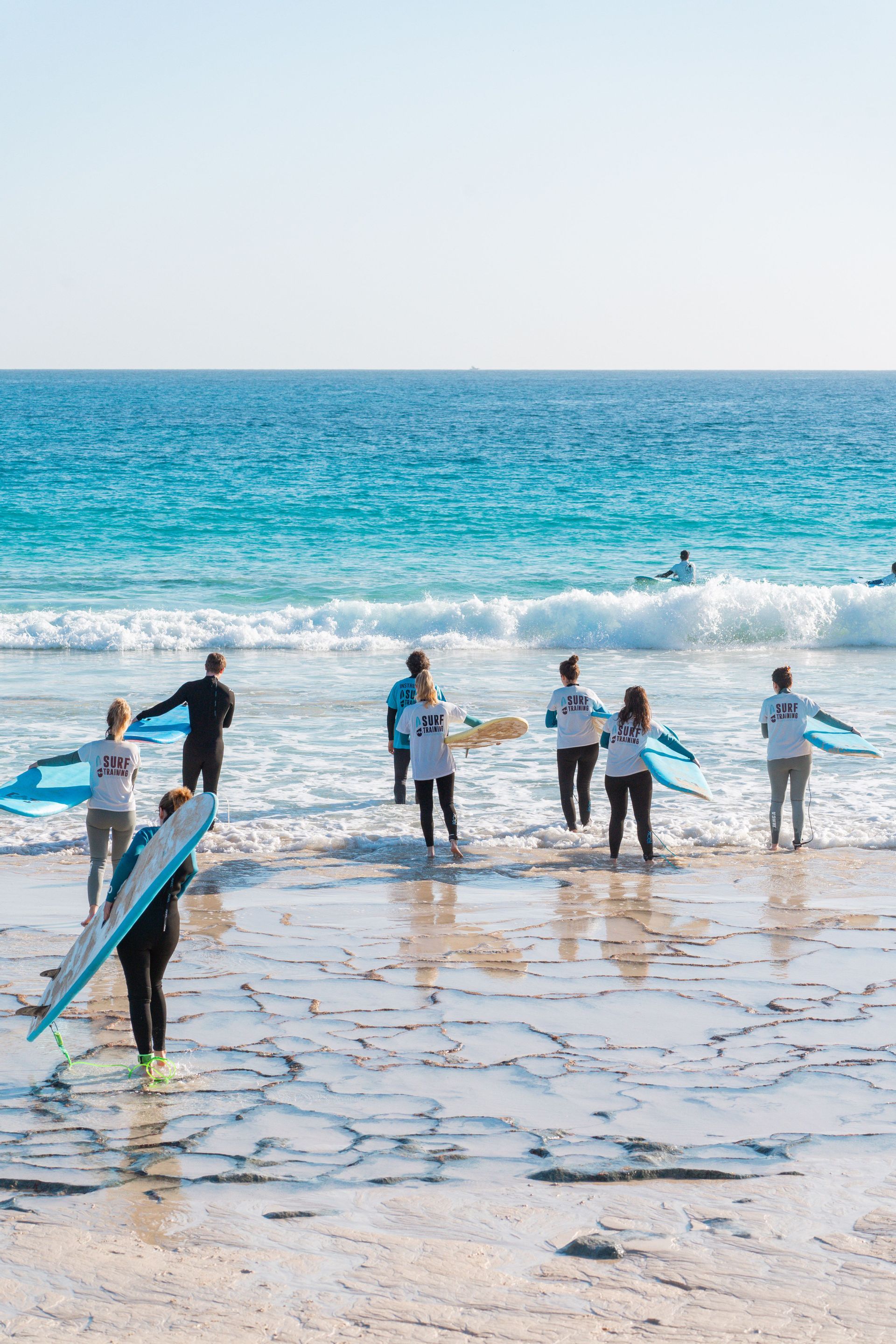 Un grupo de WeRoad con trajes de neopreno lleva tablas de surf al mar desde una costa rocosa aterrazada para una clase de surf.