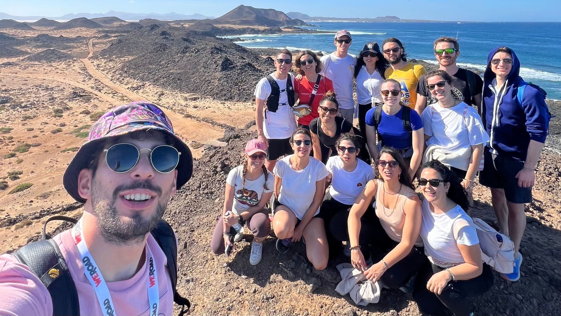Un viaje en grupo de WeRoad se toma una selfie en una colina rocosa con vistas a una costa soleada y el océano.