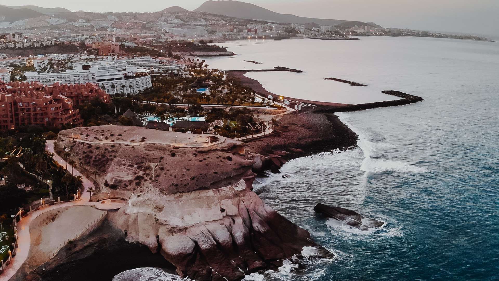 Una vista aerea di una città costiera al tramonto, con edifici lungo una costa rocciosa e spiagge di sabbia scura accanto all'oceano.