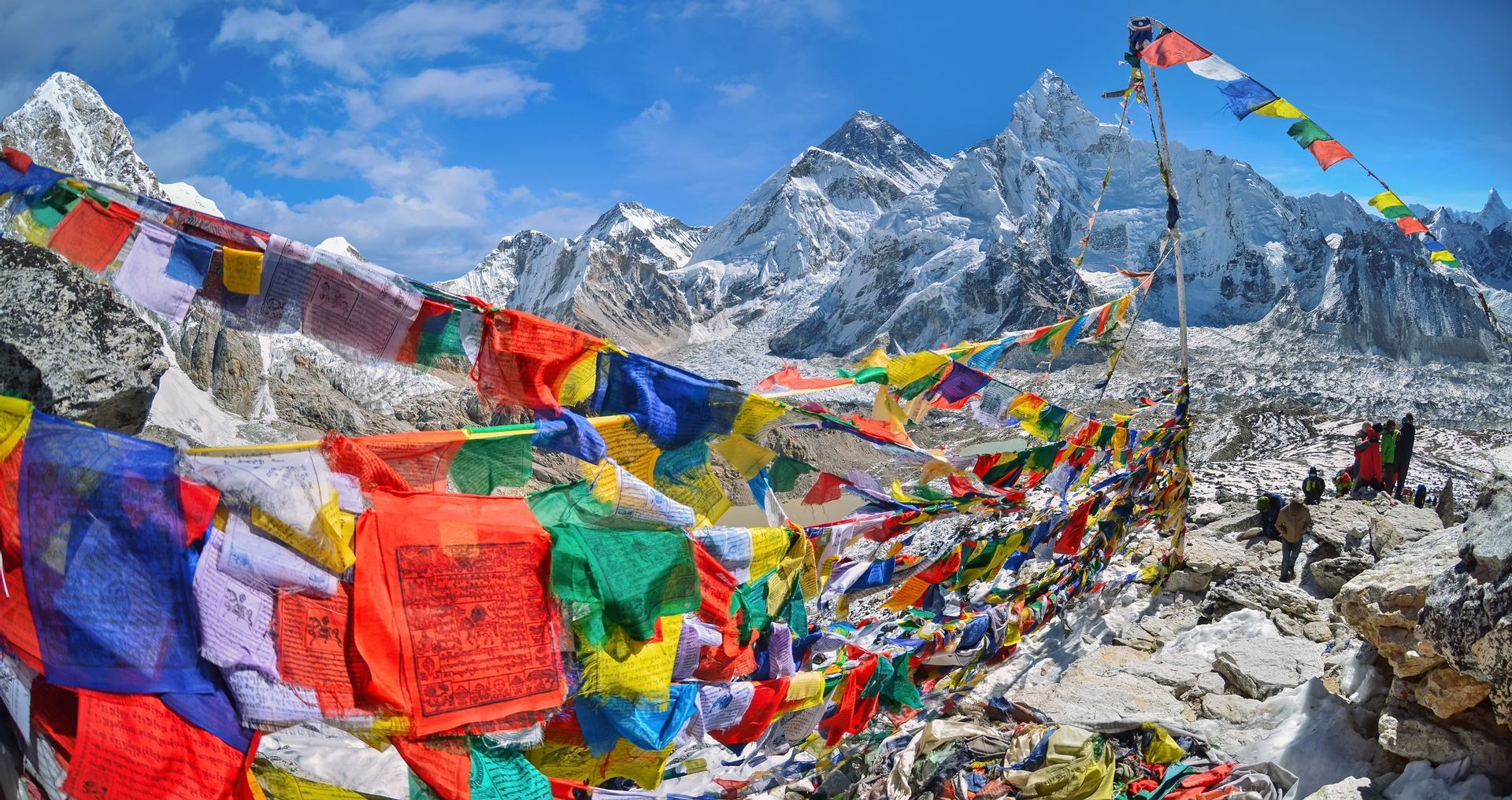 Bandiere di preghiera colorate si estendevano su un punto panoramico roccioso, con montagne innevate e un ghiacciaio sullo sfondo sotto un cielo blu.
