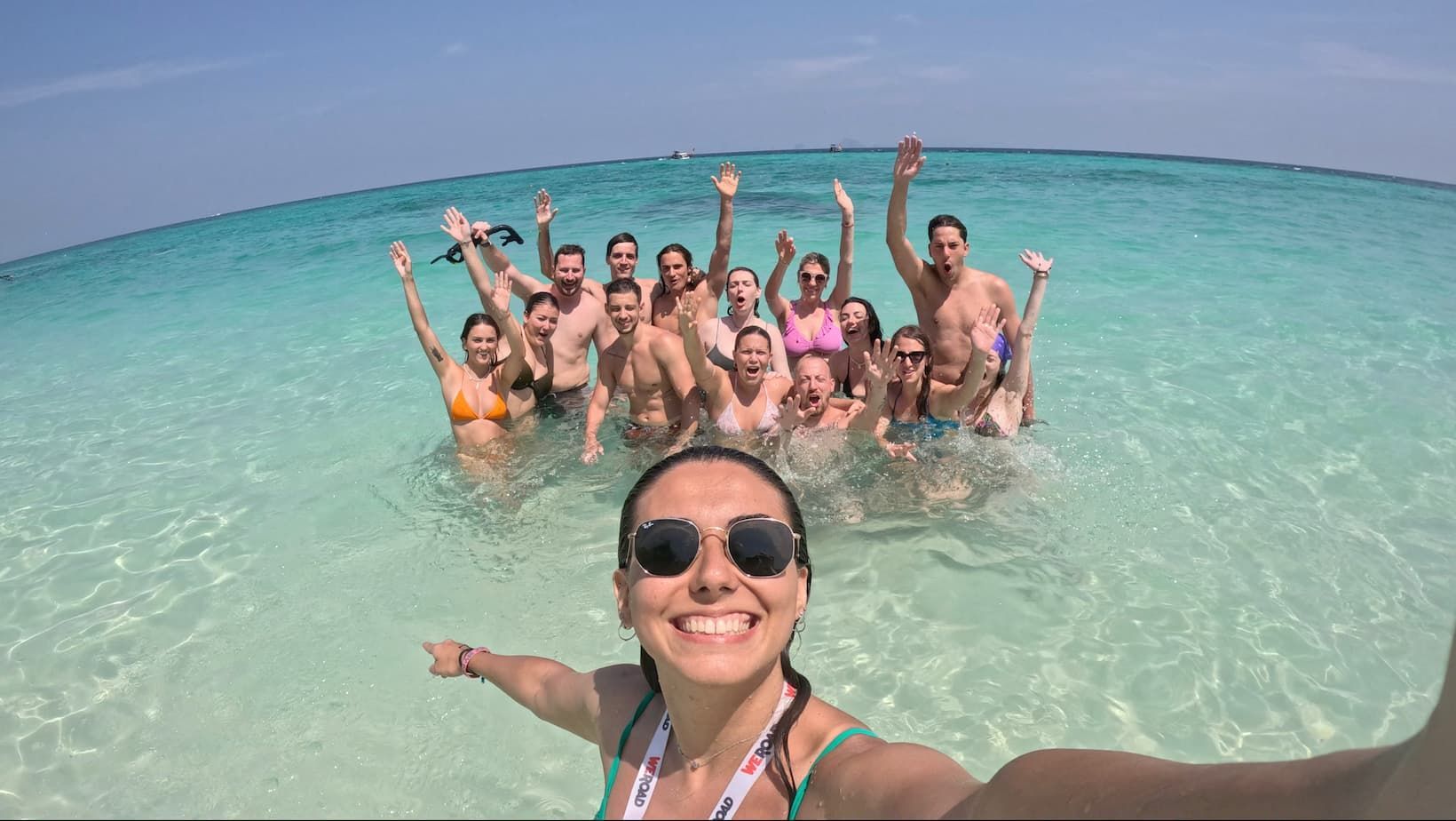 Une femme prend un selfie avec le groupe WeRoad, souriante et saluant, dans une eau de mer claire et turquoise.
