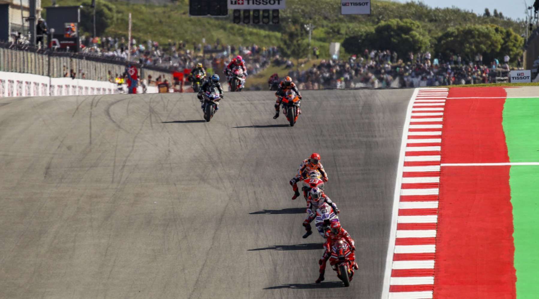 Un gruppo di motociclisti scende a tutta velocità una curva ripida su una pista professionale durante una competizione, con spettatori sullo sfondo.