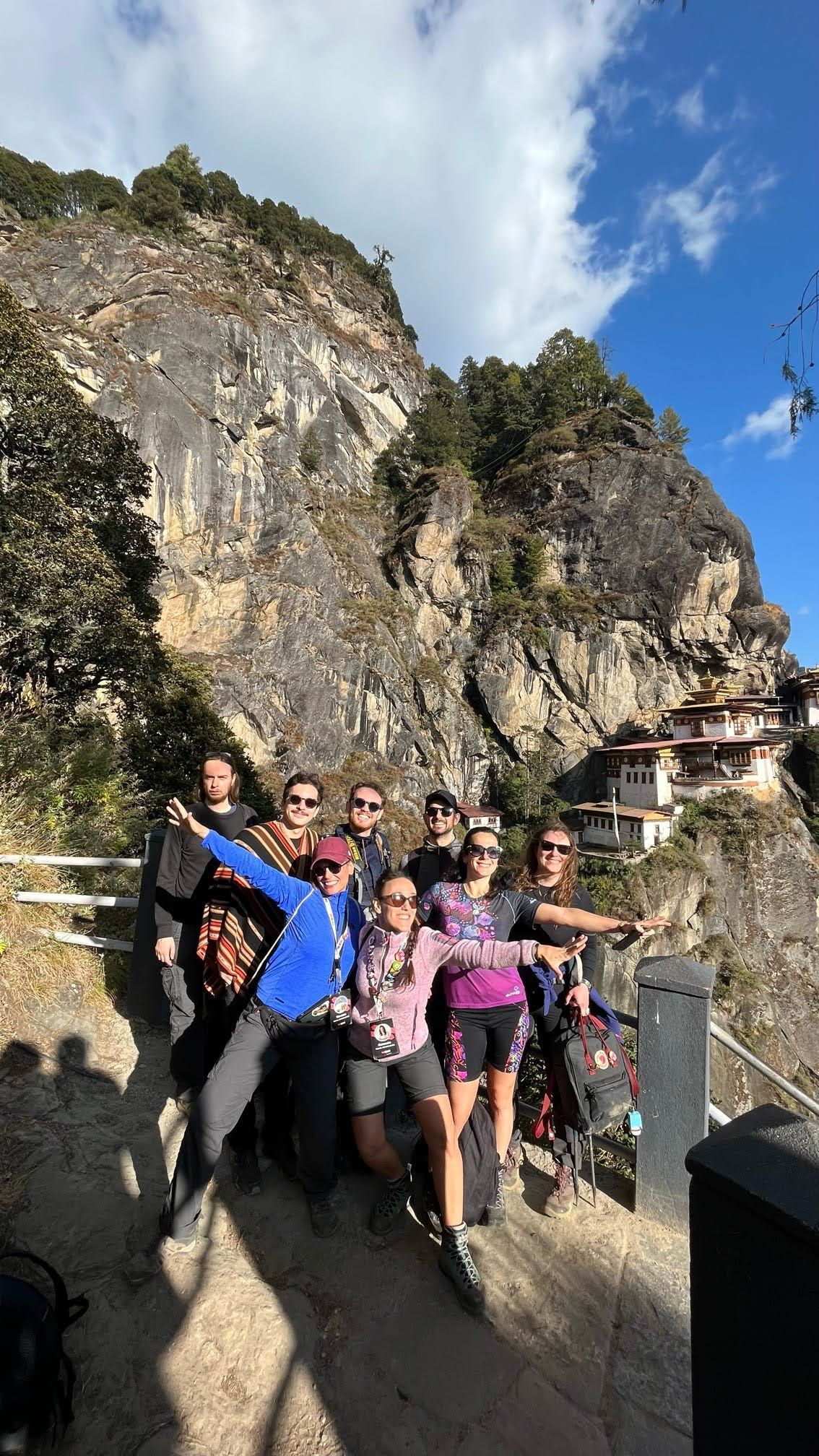 Un gruppo WeRoad in viaggio posa per una foto su un sentiero di montagna, con un monastero costruito nella parete rocciosa visibile sullo sfondo.