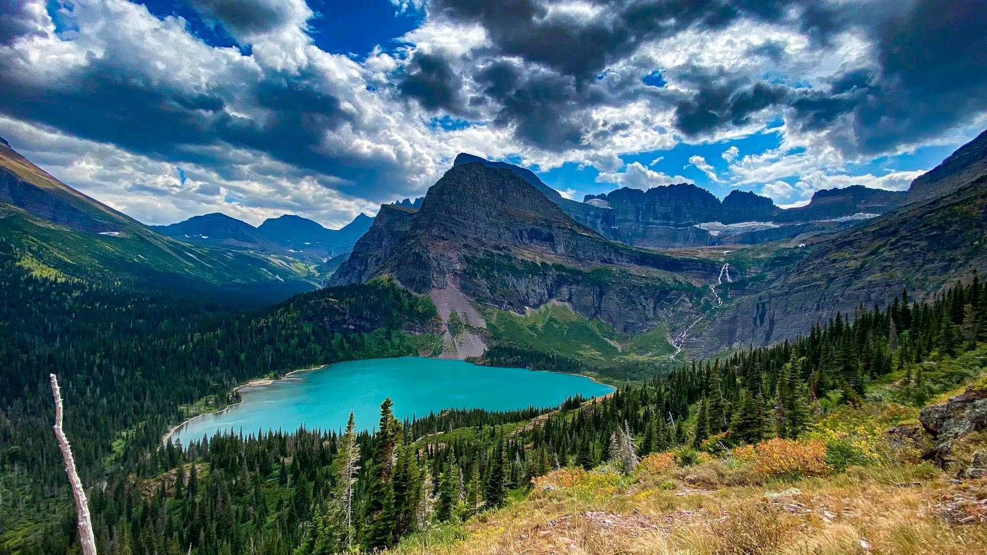 Un lago turchese brillante riposa in una valle, circondato da una fitta foresta sempreverde e aspre montagne sotto un cielo azzurro nuvoloso.