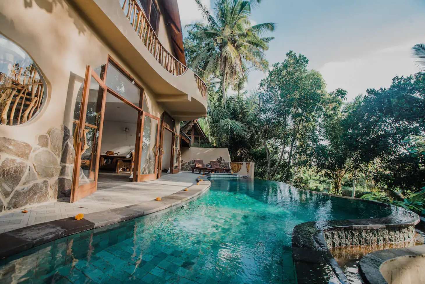 Una piscina a sfioro con acqua turchese accanto a una villa moderna con porte a vetro aperte, circondata da una rigogliosa foresta tropicale.