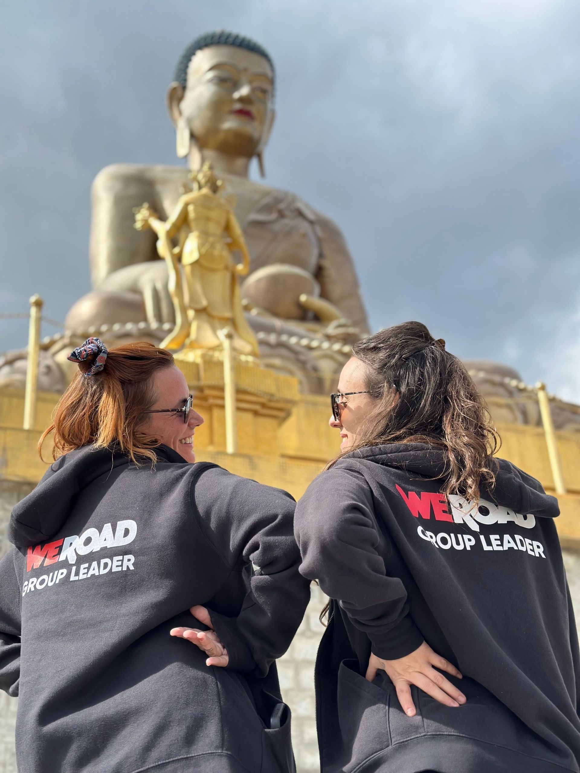Due accompagnatori WeRoad con felpe nere con cappuccio che si sorridono davanti a una gigantesca statua del Buddha dorata.