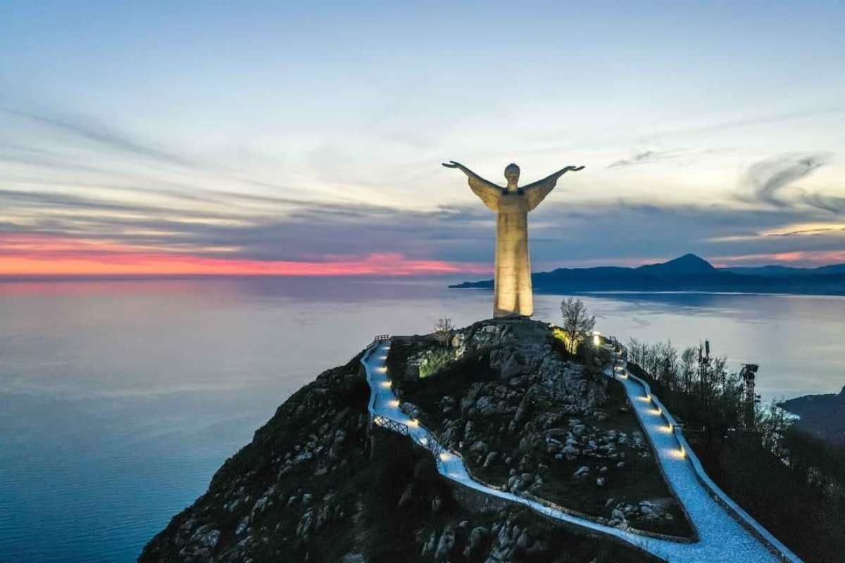 Una statua illuminata a braccia aperte si erge su una collina che domina il mare durante un tramonto colorato, con un sentiero illuminato serpeggiante.