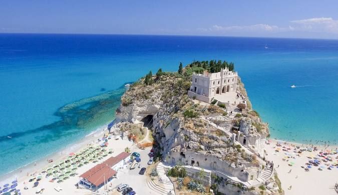 Una vista aerea di un santuario bianco su un promontorio roccioso che si affaccia su una spiaggia sabbiosa e un mare turchese sotto un cielo sereno.