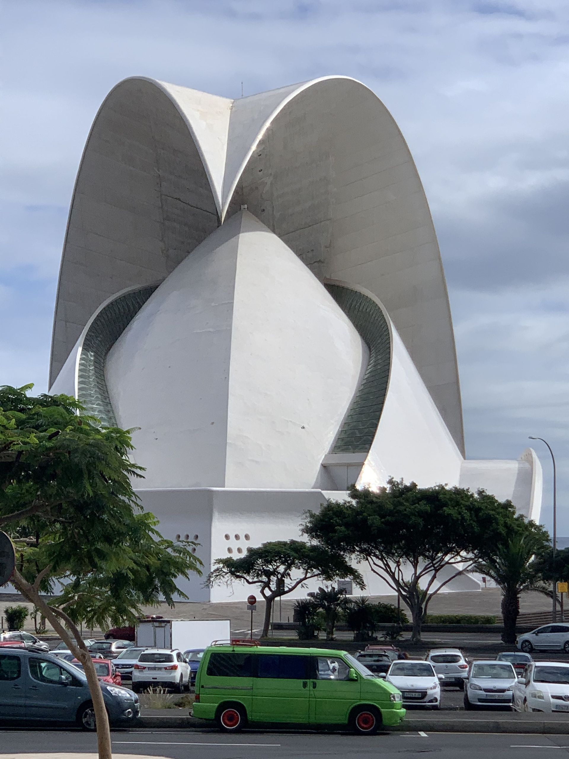 Un gran edificio blanco, con un techo ancho y curvo, se encuentra detrás de una calle bordeada de autos estacionados, incluida una camioneta verde brillante.