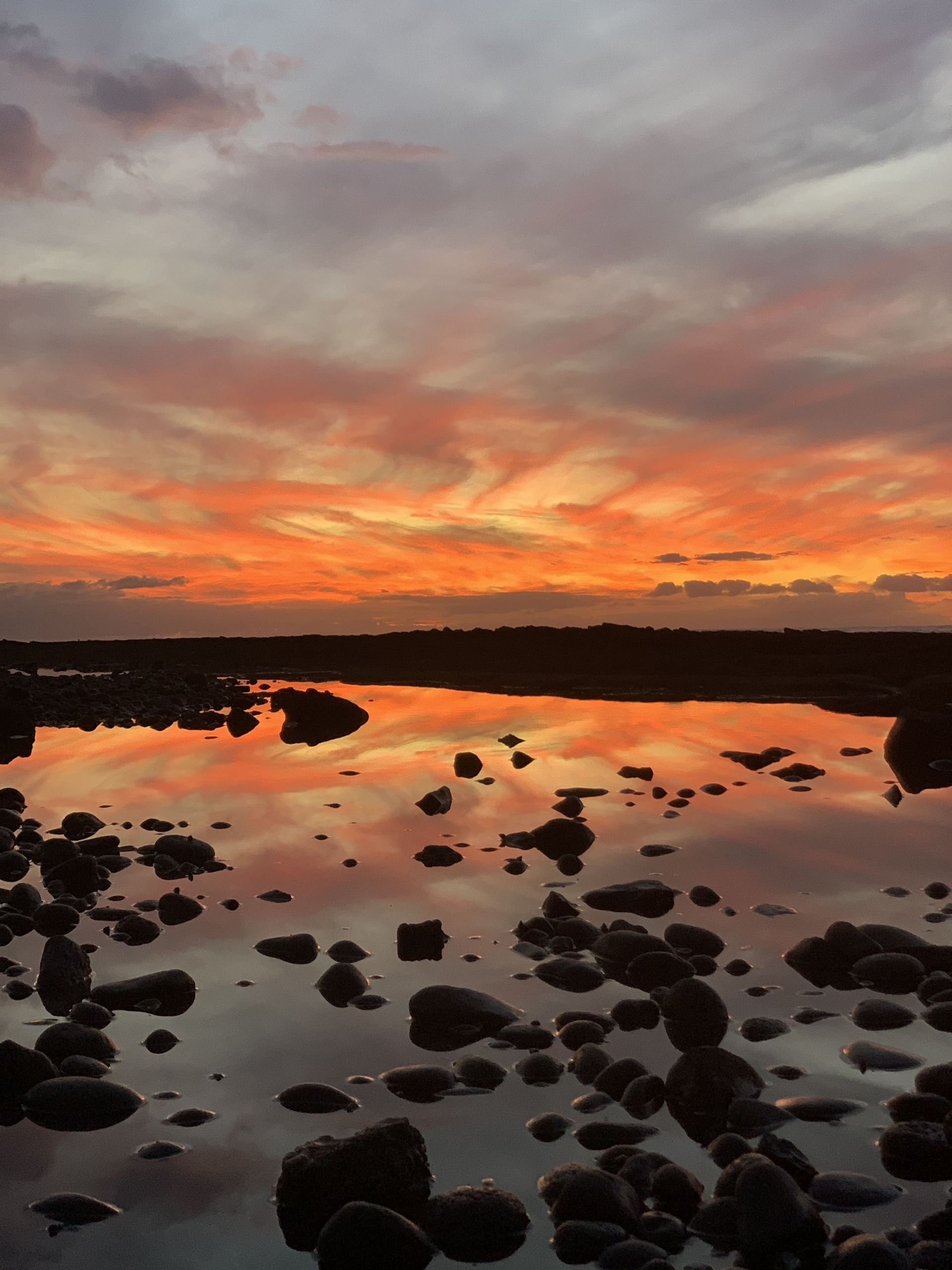 Una costa rocosa con pozas de marea que reflejan las nubes anaranjadas y rosadas de un atardecer.