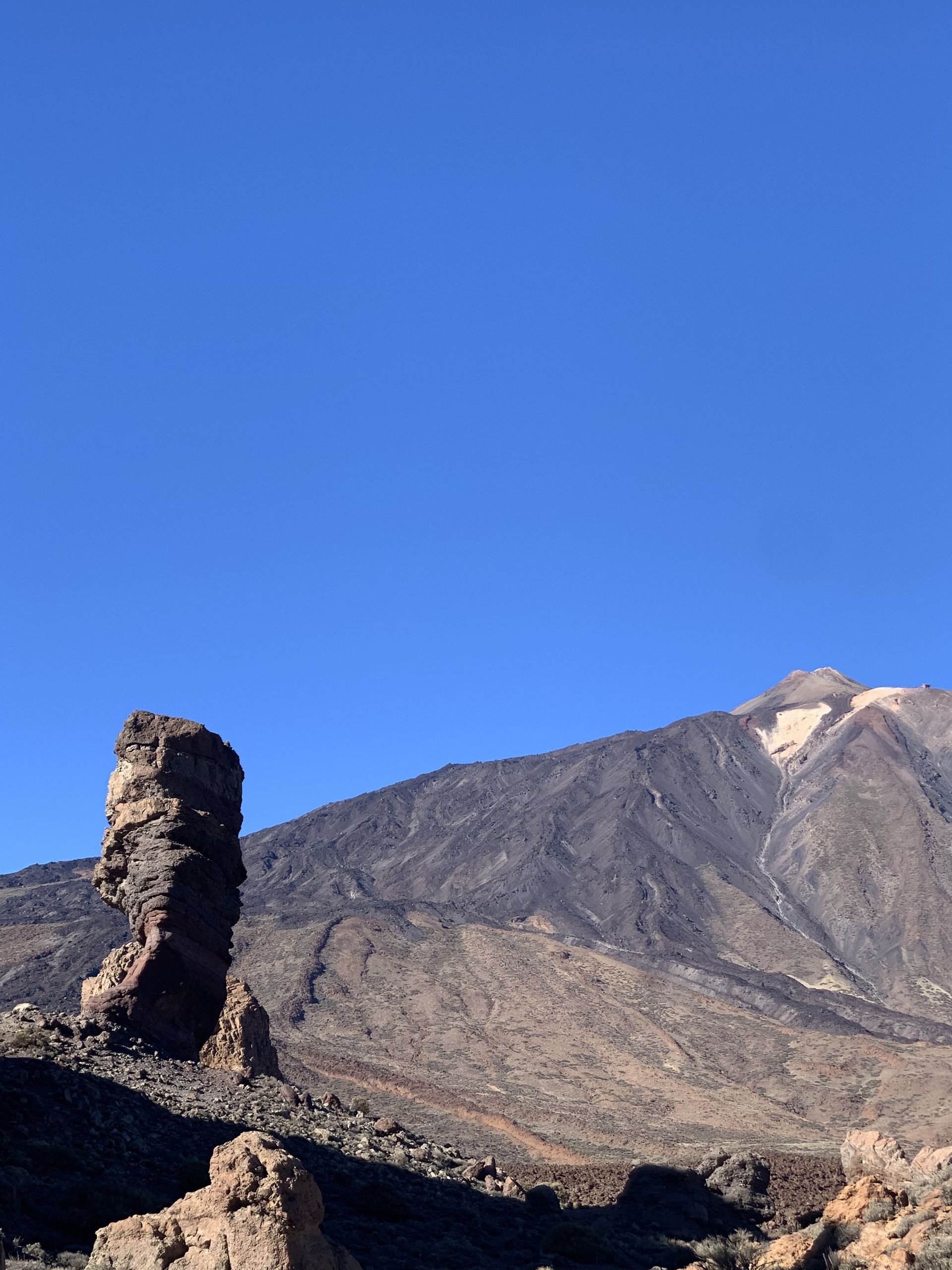 Una gran formación rocosa estratificada se eleva en un paisaje volcánico con una imponente montaña detrás bajo un cielo azul claro.