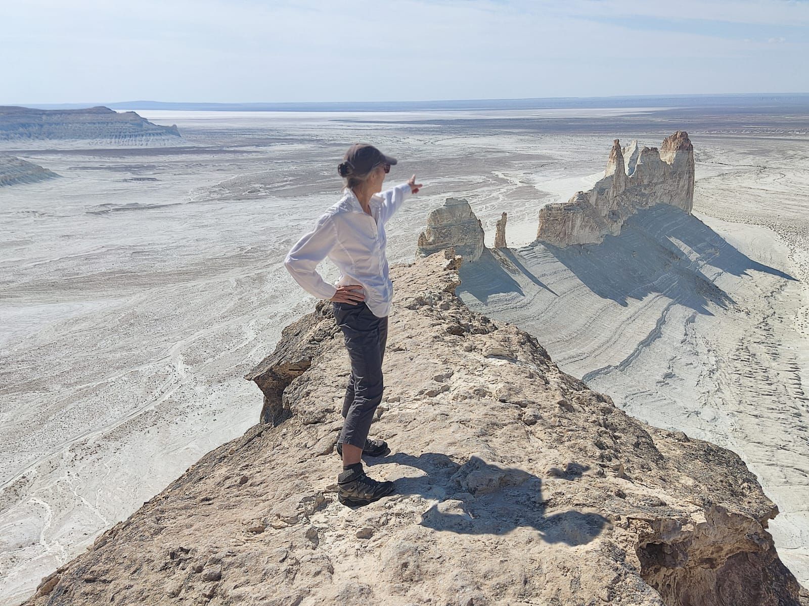 Una persona con gorra se encuentra en una estrecha cresta rocosa, señalando un vasto valle desértico soleado con formaciones rocosas blancas.