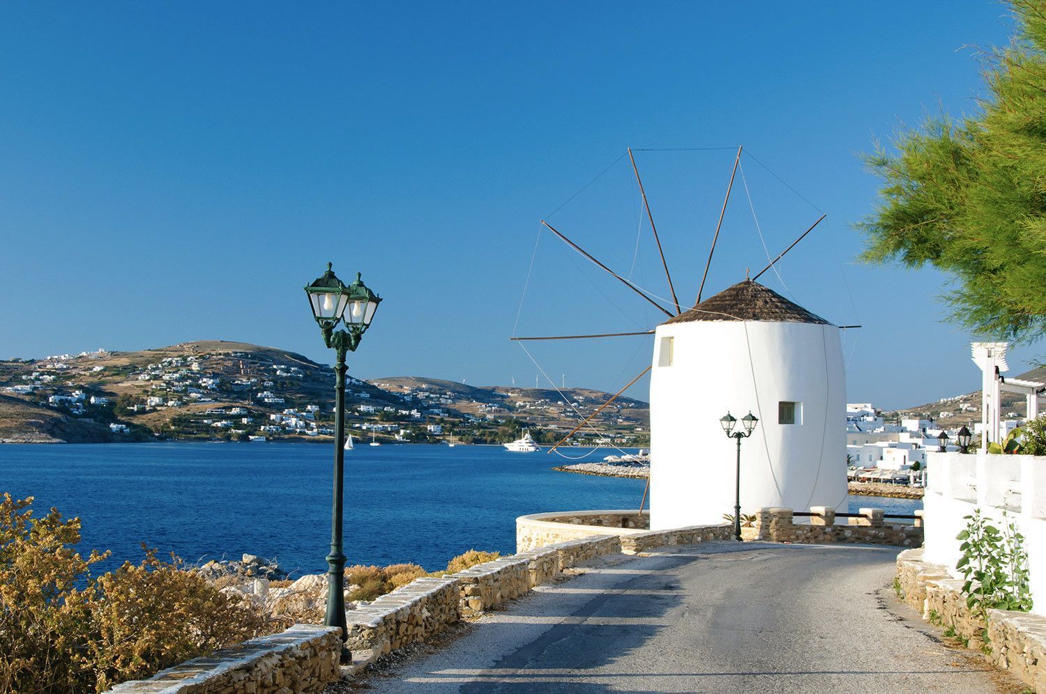 Un tradizionale mulino a vento bianco si erge su una strada costiera che domina una luminosa baia azzurra con colline e case in lontananza.