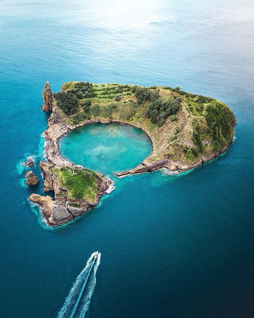 Vista aerea di un isolotto vulcanico verde con una laguna centrale turchese, mentre una motobarca passa nel mare blu profondo.
