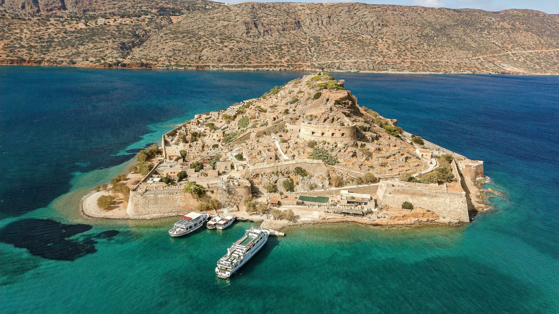 Vista aerea di un'isola storica fortificata con rovine in pietra, con diverse barche ormeggiate nell'acqua turchese circostante.