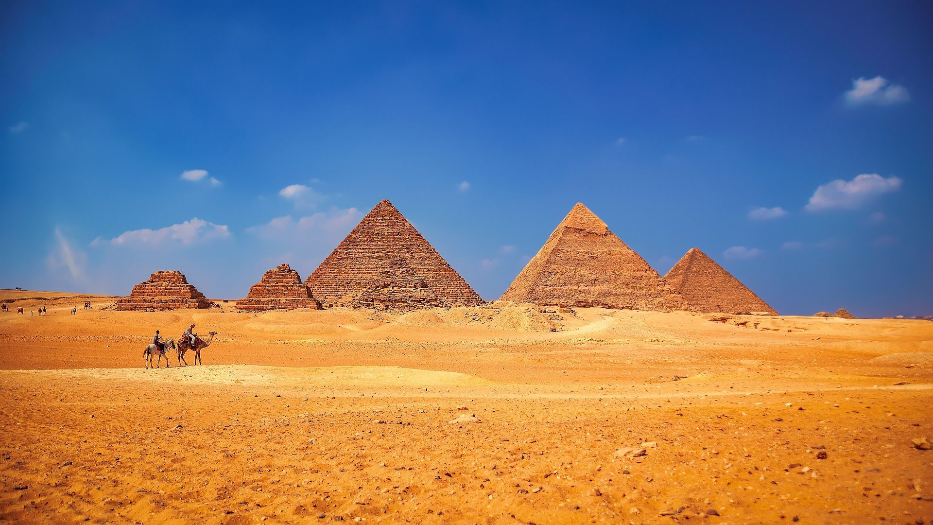 Diverse grandi piramidi si ergono dal deserto dorato sotto un cielo azzurro, con due persone su un cammello e un cavallo in primo piano.