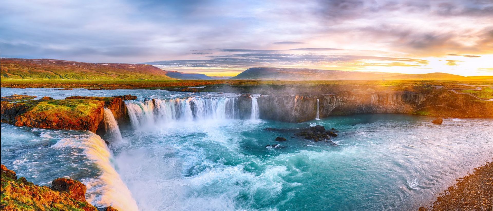 Ein breiter, mächtiger Wasserfall stürzt in einen türkisfarbenen Fluss, eingebettet in eine grüne, felsige Landschaft unter einem vom Sonnenuntergang beleuchteten Himmel.