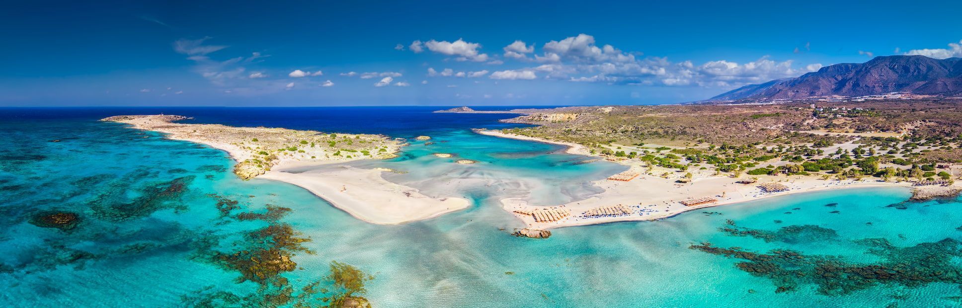 Aerial view of a sandy peninsula stretching into shallow turquoise water, with mountains on the distant coastline under a blue sky.
