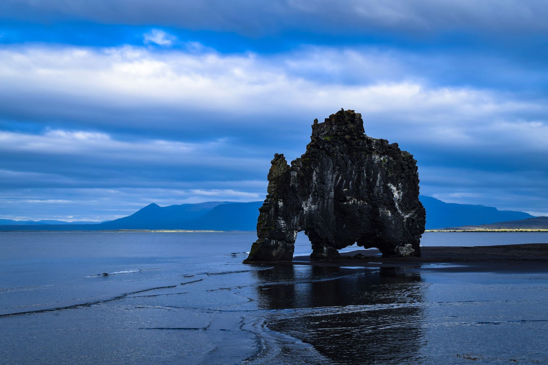 Eine große Felsformation mit einem Bogen steht im Wasser vor einem dunklen Sandstrand unter einem bewölkten blauen Himmel.