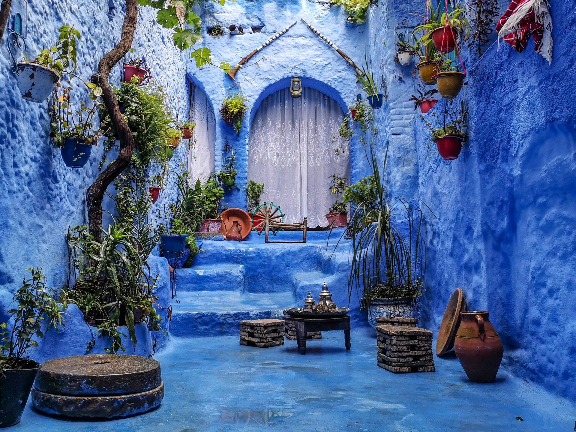 Un patio aux murs bleus texturés, avec des marches et un sol, décoré de plantes en pot, de tabourets en pierre et d'une petite table avec un service à thé.