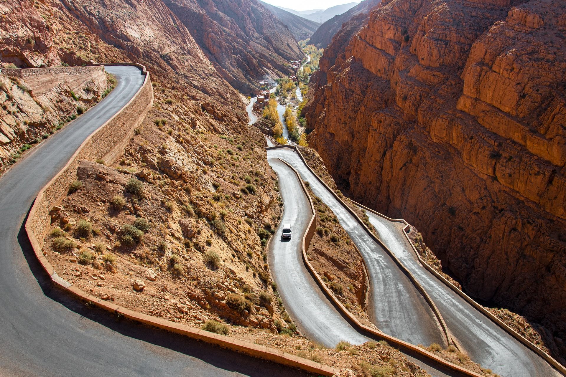 Vista aérea de un coche blanco en una carretera sinuosa con múltiples curvas cerradas, en un profundo cañón de roca roja.