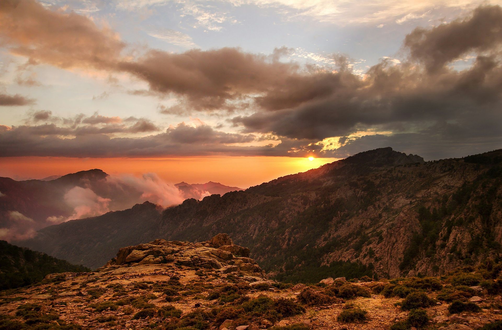Il sole tramonta dietro una catena montuosa scoscesa, proiettando un bagliore arancione su un cielo nuvoloso e sulle valli sottostanti.