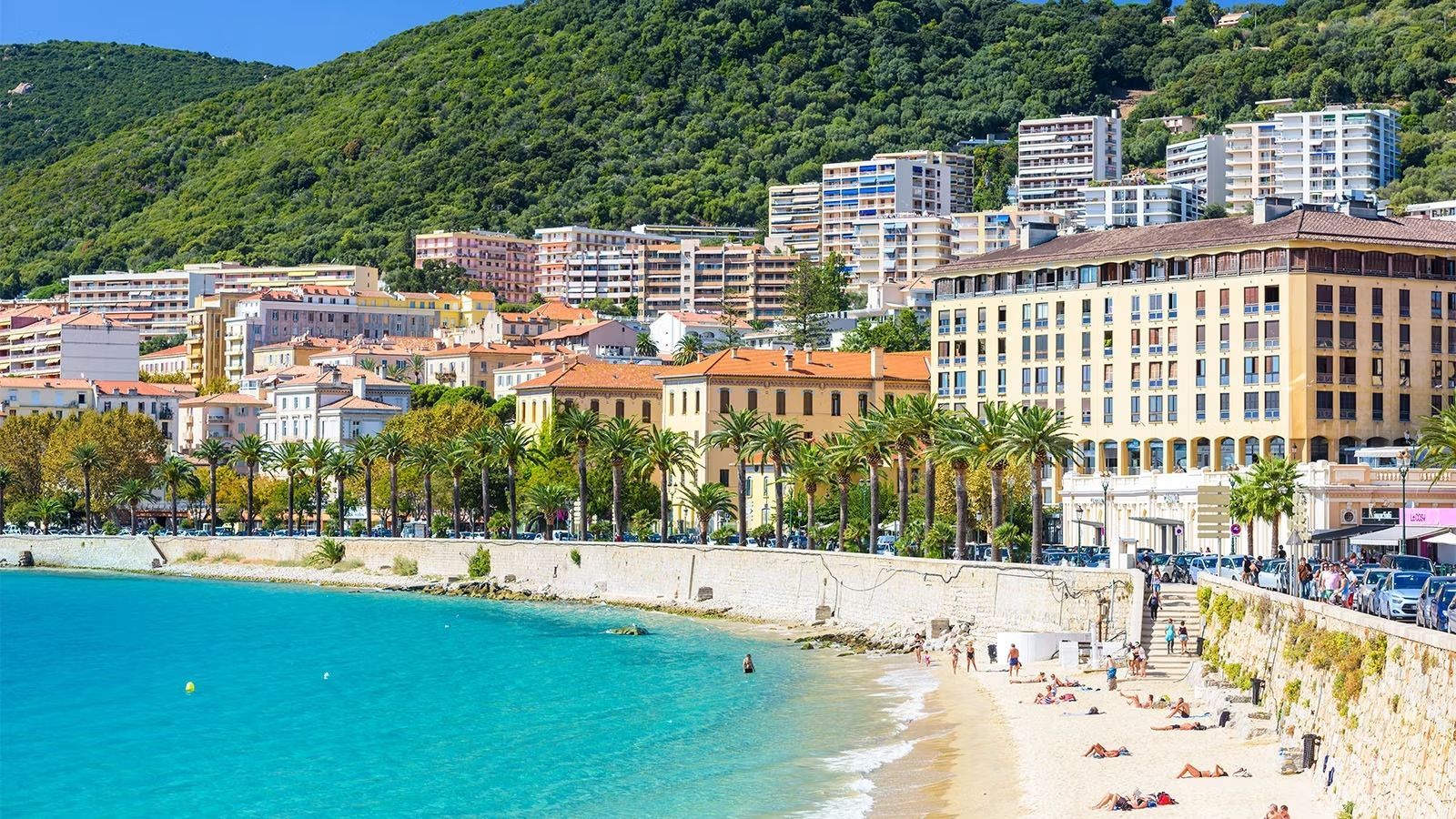Una soleggiata cittadina costiera con palme ed edifici adagiati su una collina verde, che si affaccia su una spiaggia di sabbia e un mare turchese.