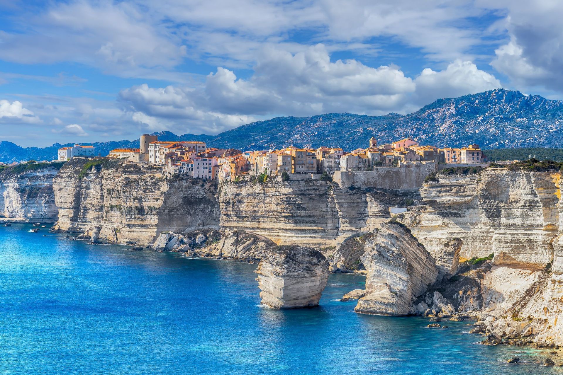 Una città costiera con edifici tradizionali si erge su alte scogliere bianche, affacciata su un mare azzurro, con montagne sullo sfondo.
