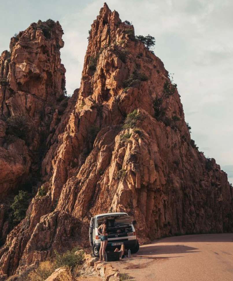 Un furgone bianco con il cofano aperto è parcheggiato su una strada di montagna ai piedi di una grande formazione rocciosa rossa, con due persone nelle vicinanze.