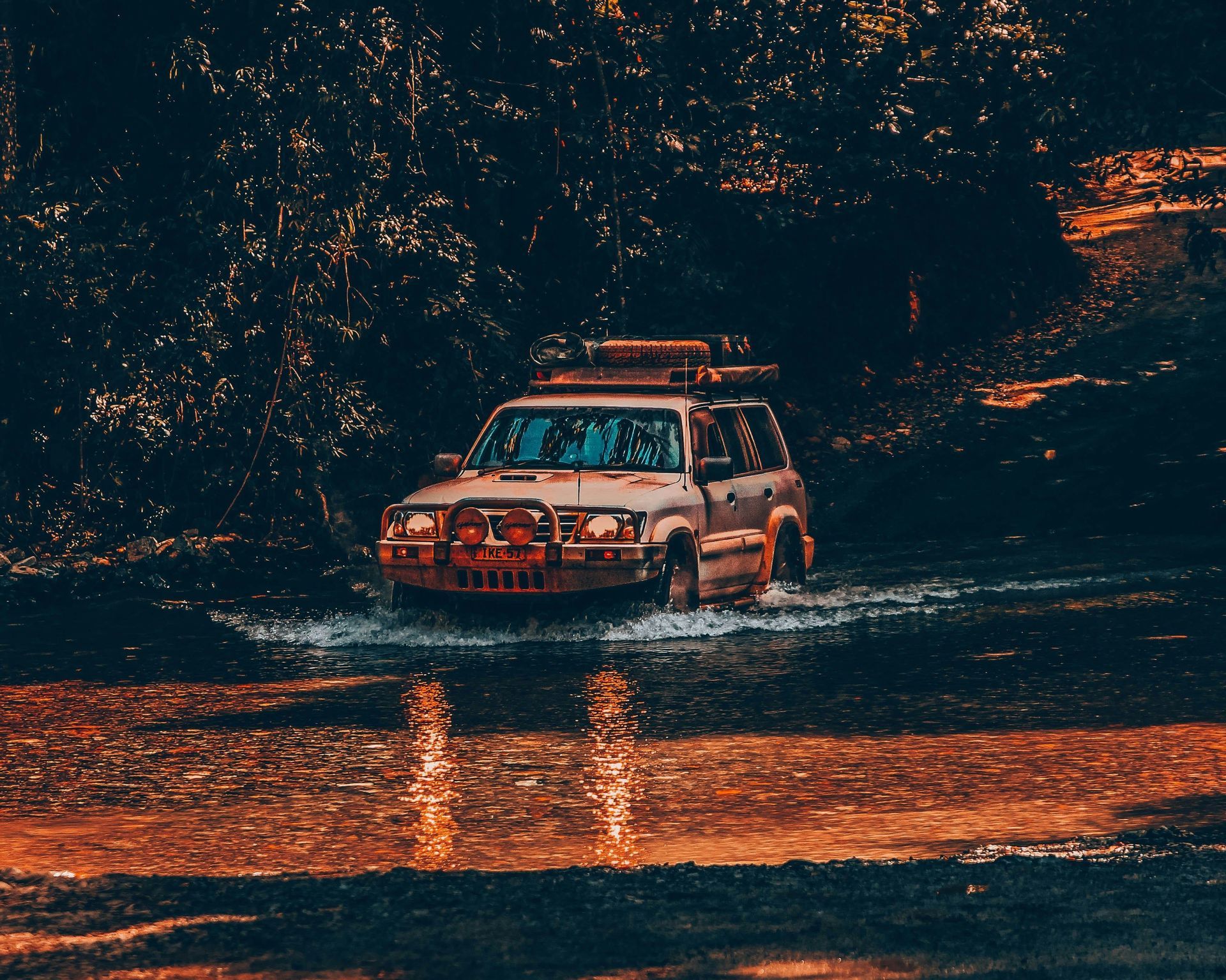 Un veicolo fuoristrada bianco attraversa un fiume poco profondo, schizzando acqua, con una fitta foresta sullo sfondo al tramonto.