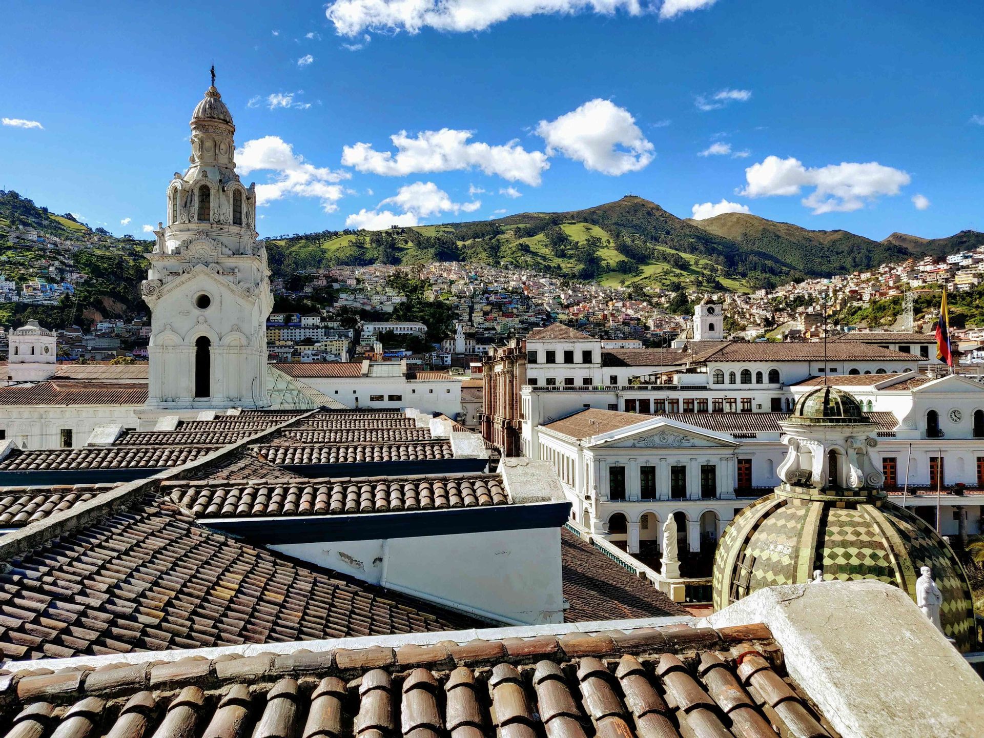 Una vista sobre los tejados de terracota de una ciudad con una torre de iglesia blanca y un edificio abovedado, con una montaña verde de fondo bajo un cielo azul.