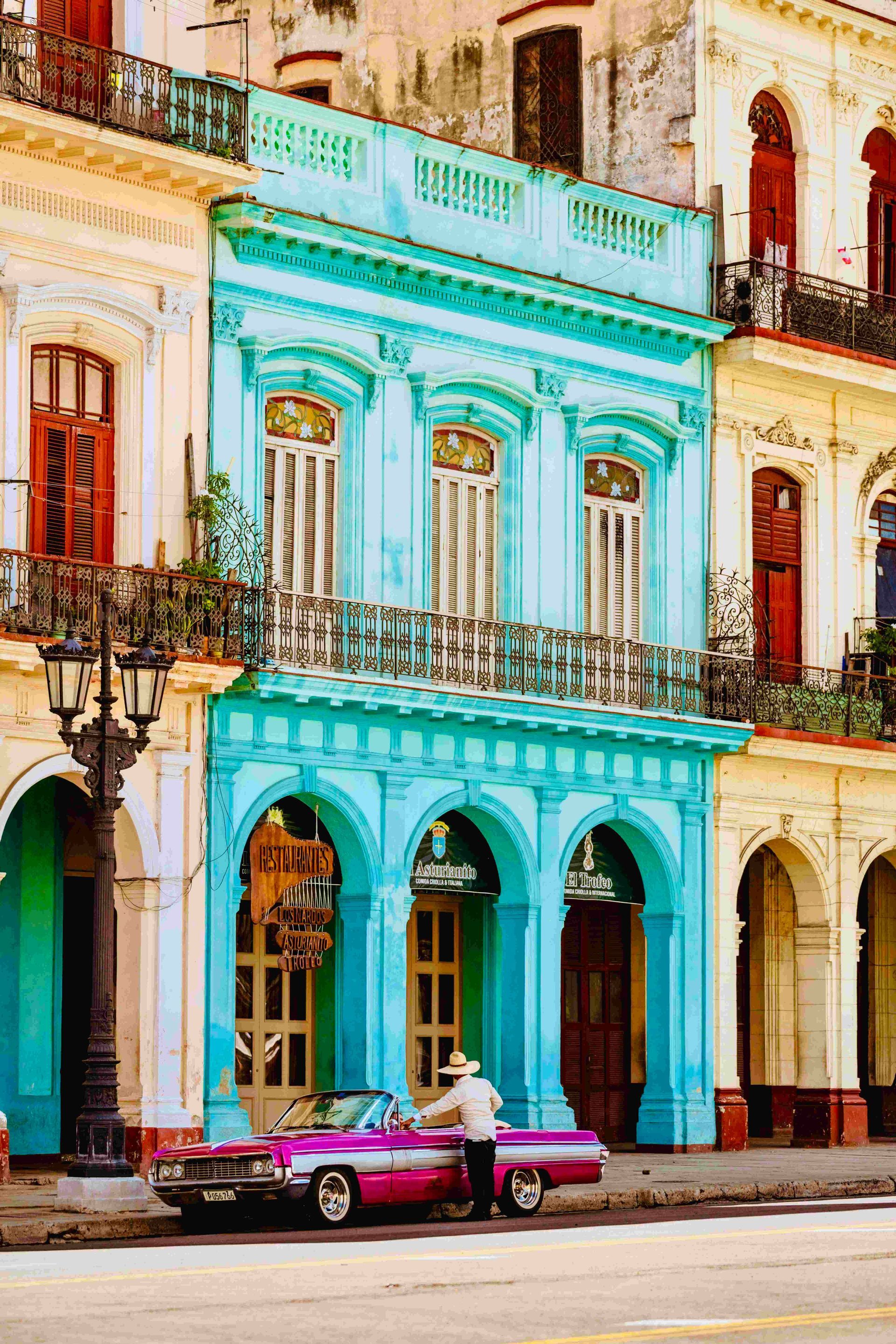 Una persona con sombrero de paja junto a un coche descapotable rosa vintage, aparcado frente a edificios coloridos y ornamentados con balcones.