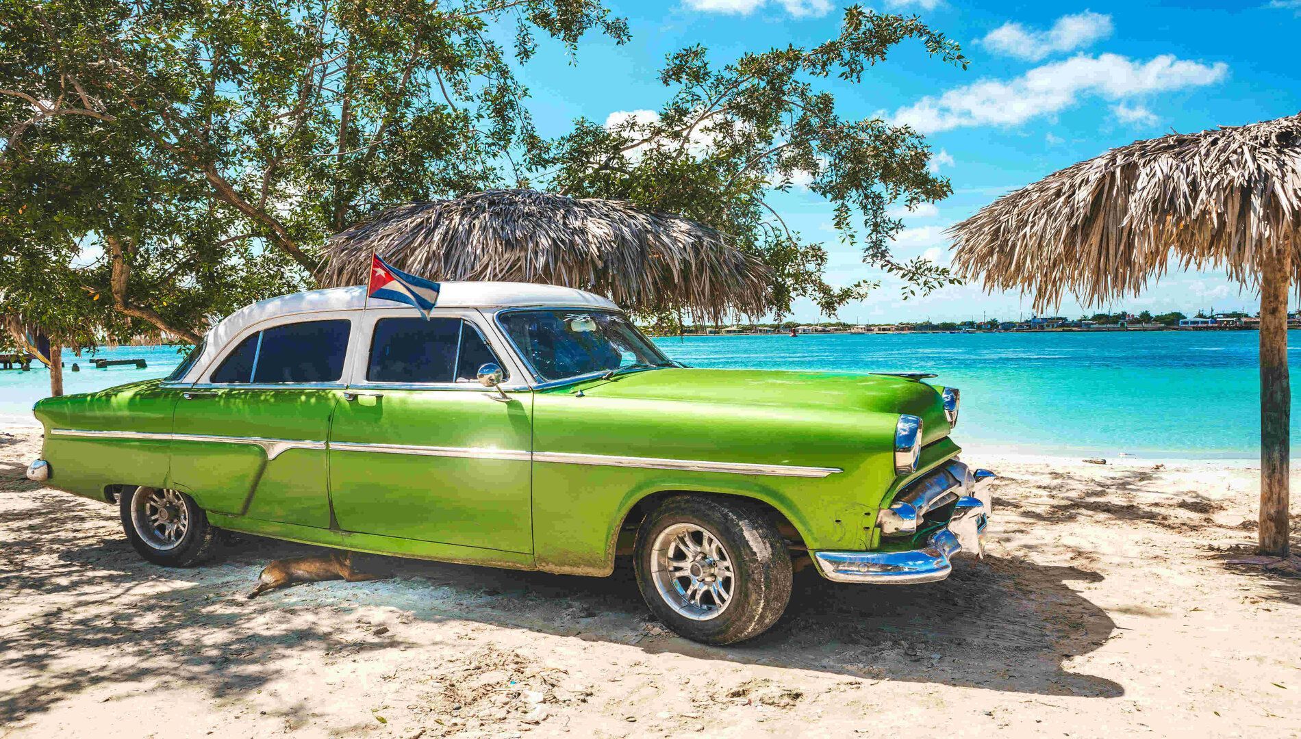 Un'auto d'epoca verde con bandiera cubana parcheggiata su una spiaggia sabbiosa accanto a un mare turchese sotto un ombrellone di paglia.