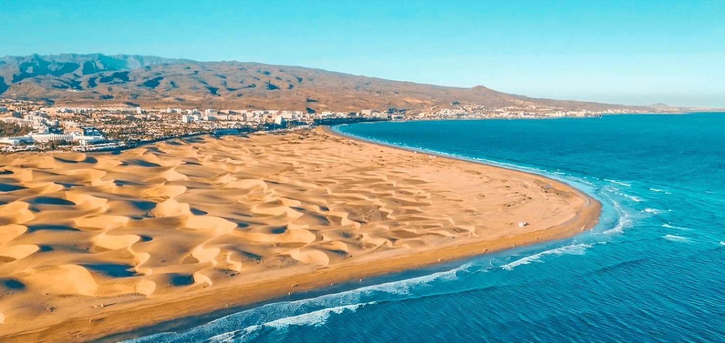 Veduta aerea di dune di sabbia dorata che incontrano un oceano blu intenso, con una città costiera e montagne sullo sfondo.