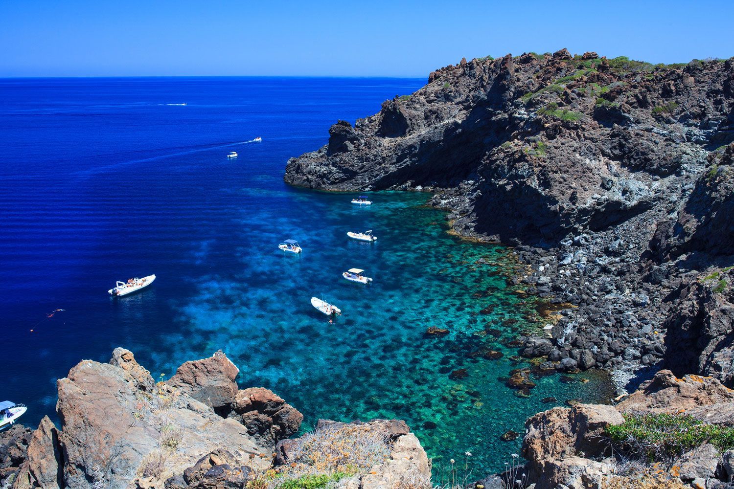 Una vista aerea di diverse barche che galleggiano su acqua limpida e turchese all'interno di una caletta rocciosa, sotto un cielo azzurro brillante.