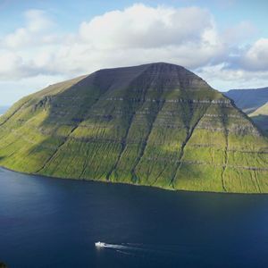 Una montagna verde conica si erge dalle acque blu profonde, con una piccola barca che naviga nelle vicinanze e altre cime in lontananza.