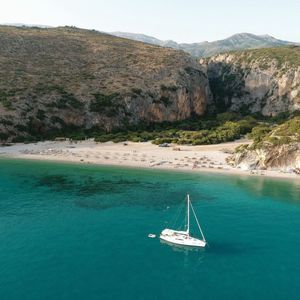 Veduta aerea di una barca a vela bianca ancorata in una cala turchese, con una spiaggia sabbiosa incastonata tra scogliere vegetate.