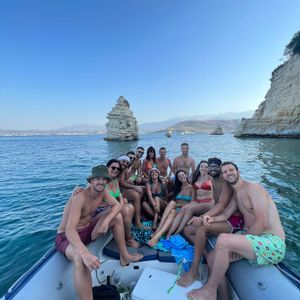 Un grupo sonriente de WeRoad en traje de baño posa en un barco en el mar, con grandes formaciones rocosas y acantilados costeros detrás.