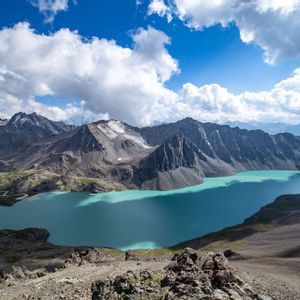 Ein Luftbild eines türkisfarbenen alpinen Sees, eingebettet in eine felsige Bergkette mit schneebedeckten Gipfeln unter einem blauen, bewölkten Himmel.