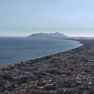 Veduta aerea di una città costiera su una baia curva, con una grande montagna oltre il mare sotto un cielo sereno.