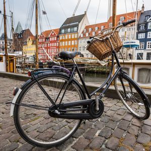 Ein schwarzes Fahrrad mit einem Weidenkorb steht auf einer Kopfsteinpflasterstraße neben einem Kanal mit Booten und bunten Gebäuden.