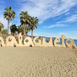Una grande scultura di sabbia che forma la scritta 'Malagueta' su una spiaggia soleggiata, con palme e il mare sullo sfondo.