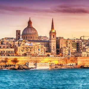 Una città costiera con una grande cupola e un campanile è illuminata lungo il bordo dell'acqua sotto un cielo al tramonto colorato.