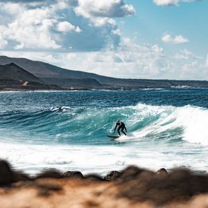 Un surfista in muta nera cavalca un'onda turchese, con una costa rocciosa e montuosa sullo sfondo.
