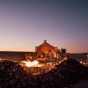 A campfire burns in a rock-lined pit in front of a large, illuminated tent in a desert campsite at dusk.