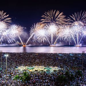 Una grande folla di persone radunata su una spiaggia osserva un enorme spettacolo di fuochi d'artificio che esplode sul mare di notte.